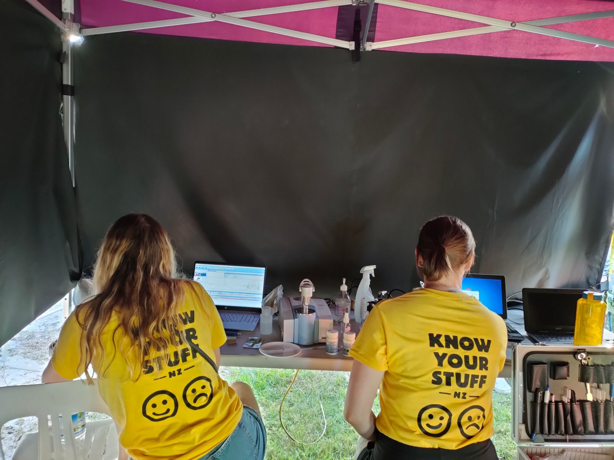 Two people at a table, with their backs to us. It&#39;s obvious they are under a gazebo in a field. There are laptops, a spectrometer, and various sciency things on the table. Both are in bright yellow &#39;Know Your Stuff&#39; tees. 